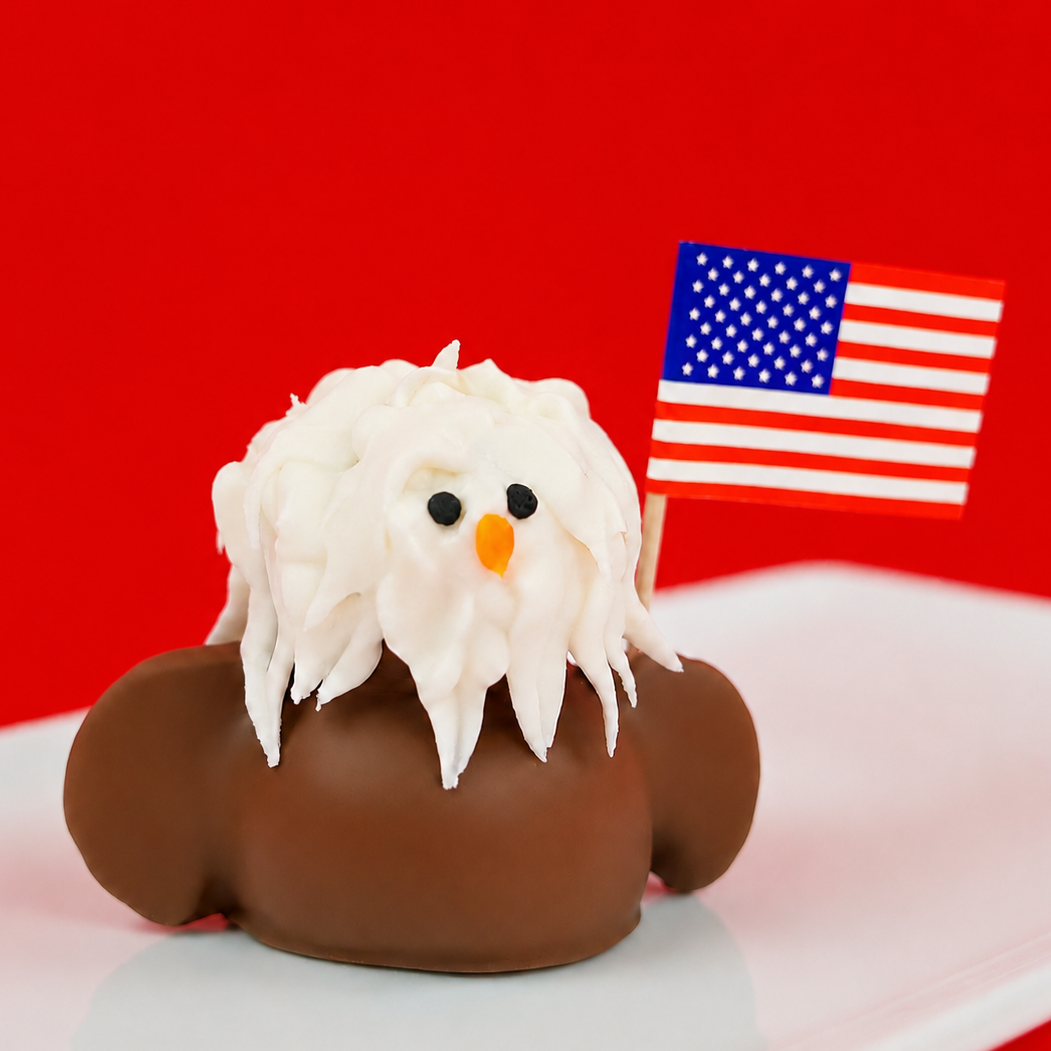 Handcrafted milk chocolate ganache bald eagle with white royal icing feather crown black eyes and orange beak and miniature American flag on a white plate with red background, extraordinary patriotic gift by Nibbles and Bits Scranton PA.