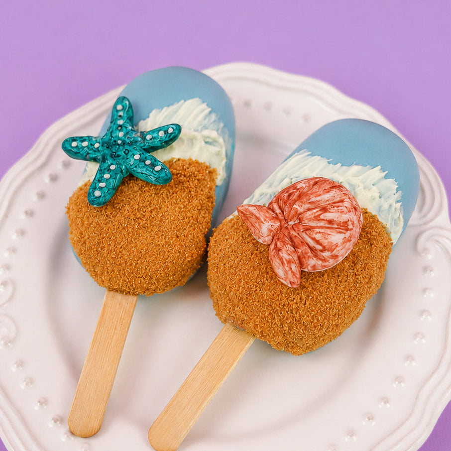 Two beach peanut butter ganache pops on a white decorative plate with lavender background, each featuring ocean blue white chocolate upper half with painted white royal icing waves and golden Biscoff cookie sand lower half, one topped with a teal hand-painted fondant starfish and one with a coral fondant seashell, handmade summer gift by Nibbles and Bits Scranton PA.