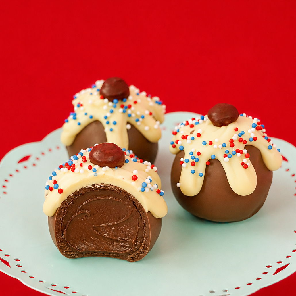A patriotic sundae milk chocolate ganache truffle revealing a rich creamy milk chocolate ganache center alongside two whole decorated truffles on a mint green cake stand, handcrafted patriotic gift by Nibbles and Bits Scranton PA.