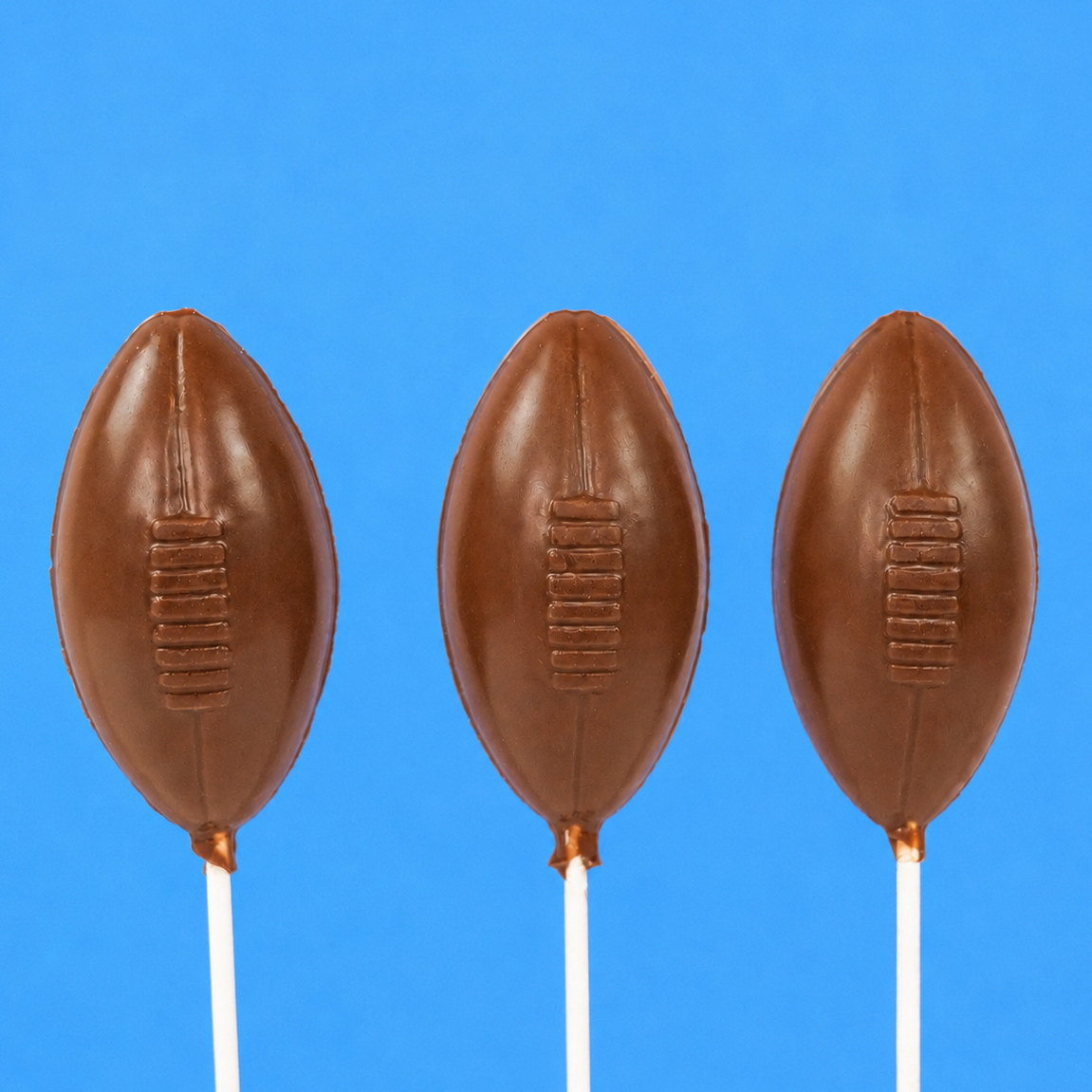 Three solid milk chocolate football shaped lollipops with molded lace stitching detail on white sticks against a blue background, handmade game day treat by Nibbles and Bits Scranton PA.