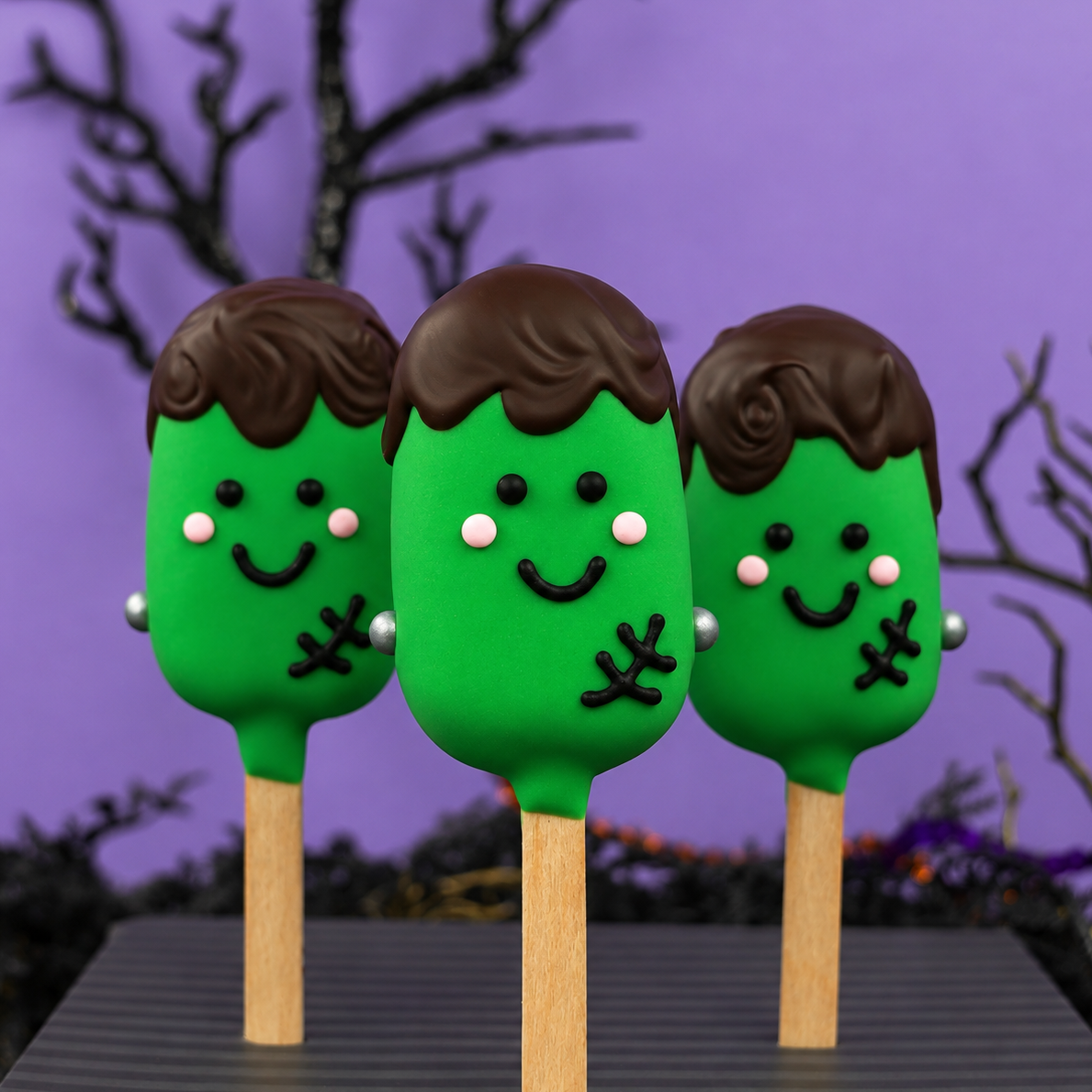 Three cookie dough pops dipped in green chocolate and decorated with Frankenstein faces on a purple Halloween background.