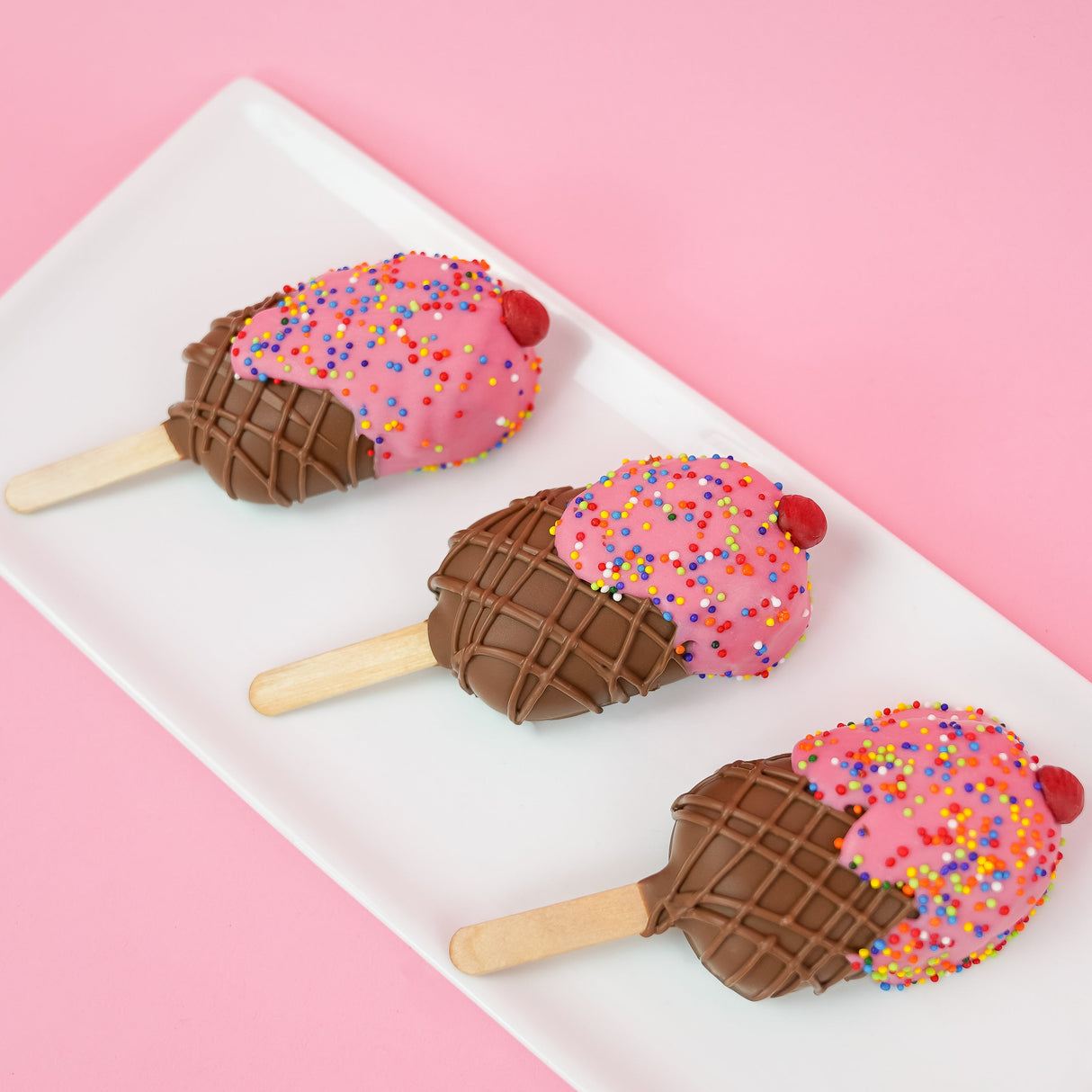 Three ice cream milk chocolate peanut butter pops on a white rectangular plate with pink background, each featuring a pink chocolate scoop with rainbow nonpareils and chocolate cherry over a crosshatch milk chocolate cone base on a stick, handmade summer treat by Nibbles and Bits Scranton PA.