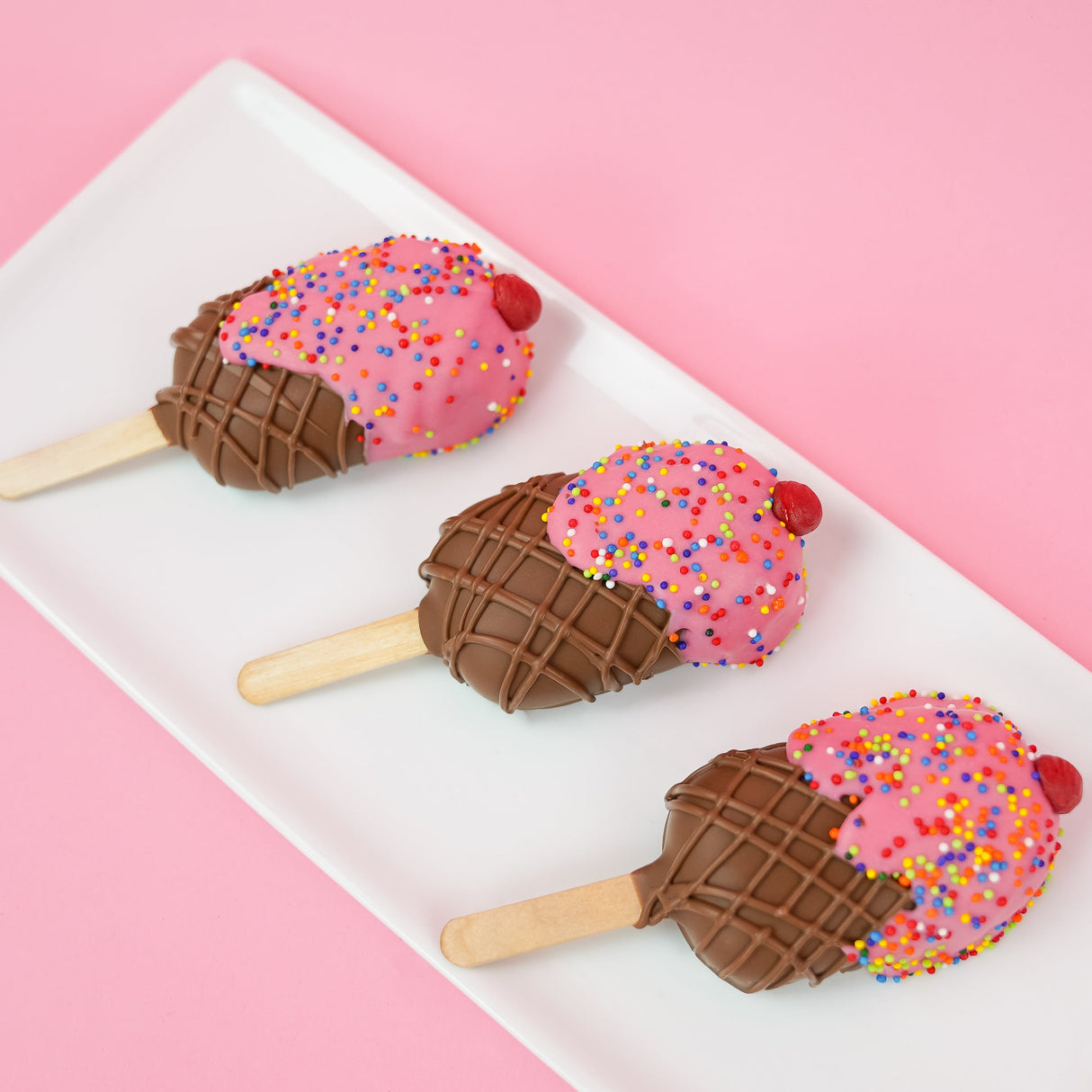 Three ice cream milk chocolate peanut butter pops on a white rectangular plate with pink background, each featuring a pink chocolate scoop with rainbow nonpareils and icing cherry over a crosshatch milk chocolate cone base on a stick, handmade summer treat by Nibbles and Bits Scranton PA.