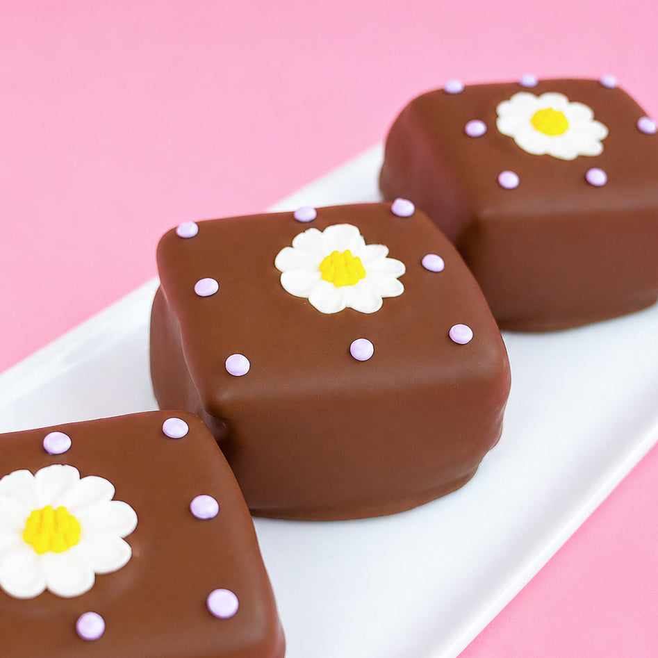 Three milk chocolate daisy s'mores on a white rectangular plate with pink background, each decorated with a white fondant daisy with yellow center and soft lavender icing dots, handmade spring treat by Nibbles and Bits Scranton PA.