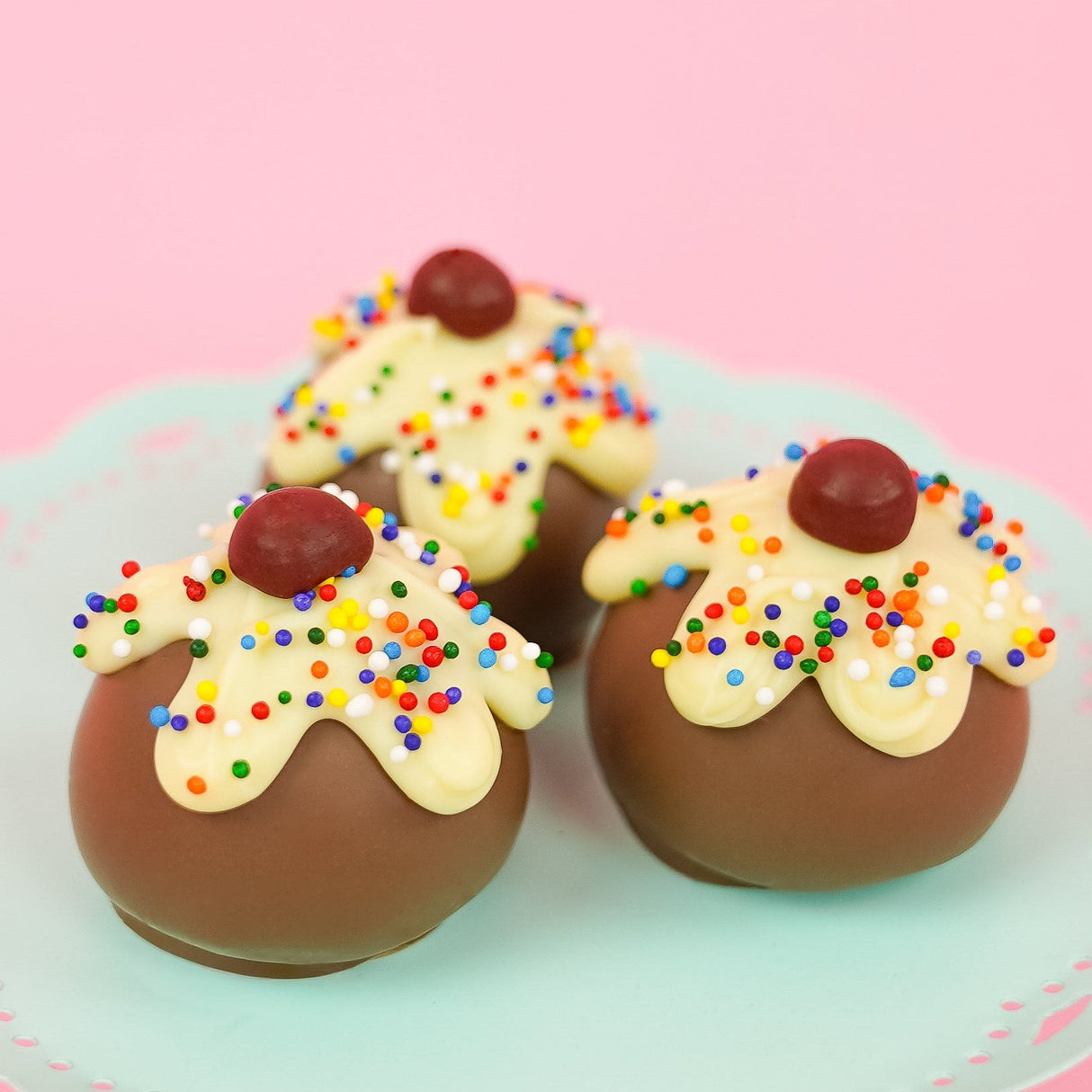 Three milk chocolate ganache sundae truffles on a mint green cake stand with pink background, each topped with dripping white chocolate rainbow nonpareil sprinkles and a cherry, handmade celebration gift by Nibbles and Bits Scranton PA.