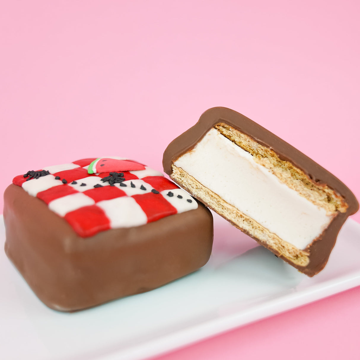A milk chocolate picnic blanket s'more revealing thick homemade vanilla marshmallow layer and golden graham cracker base alongside a whole decorated s'more on a plate with pink background, handcrafted summer treat by Nibbles and Bits Scranton PA.