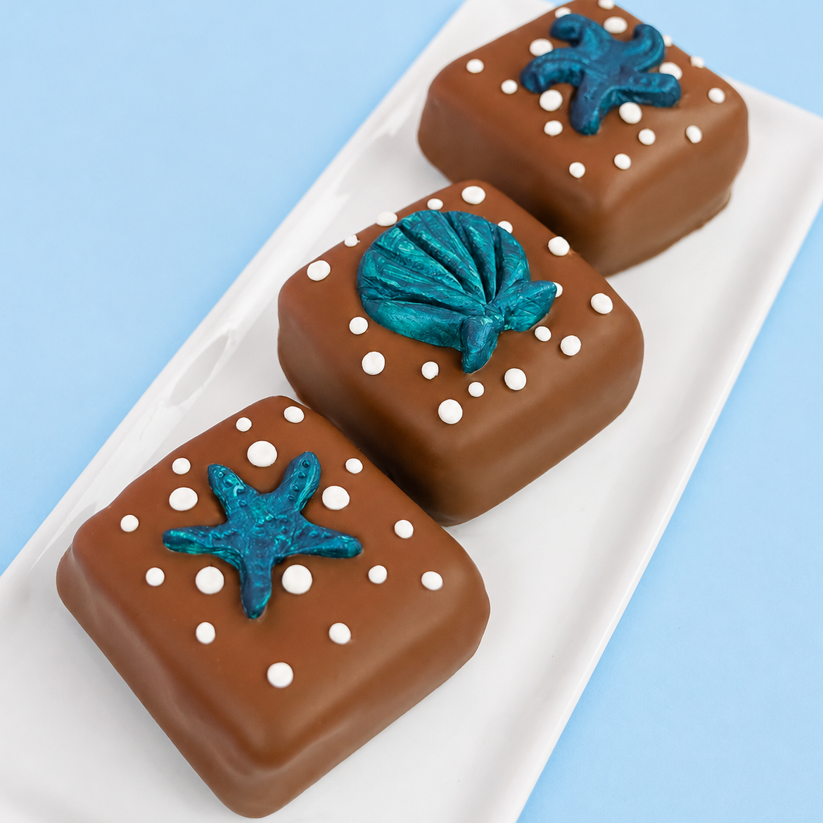 Three milk chocolate sea shell s'mores on a white rectangular plate with blue background, each decorated with a hand-painted metallic teal fondant ocean creature including a starfish a scallop shell and a sea creature surrounded by white bubble dot details, handmade coastal gift by Nibbles and Bits Scranton PA.
