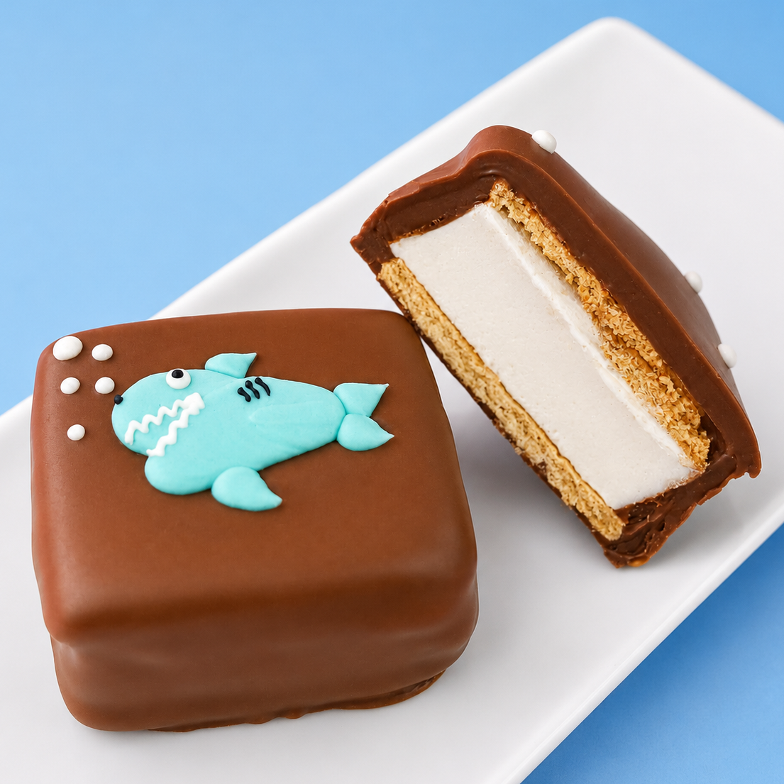 A milk chocolate shark s'more revealing thick homemade vanilla marshmallow layer and golden graham cracker alongside a whole decorated s'more on a white plate with blue background, handcrafted ocean treat by Nibbles and Bits Scranton PA.