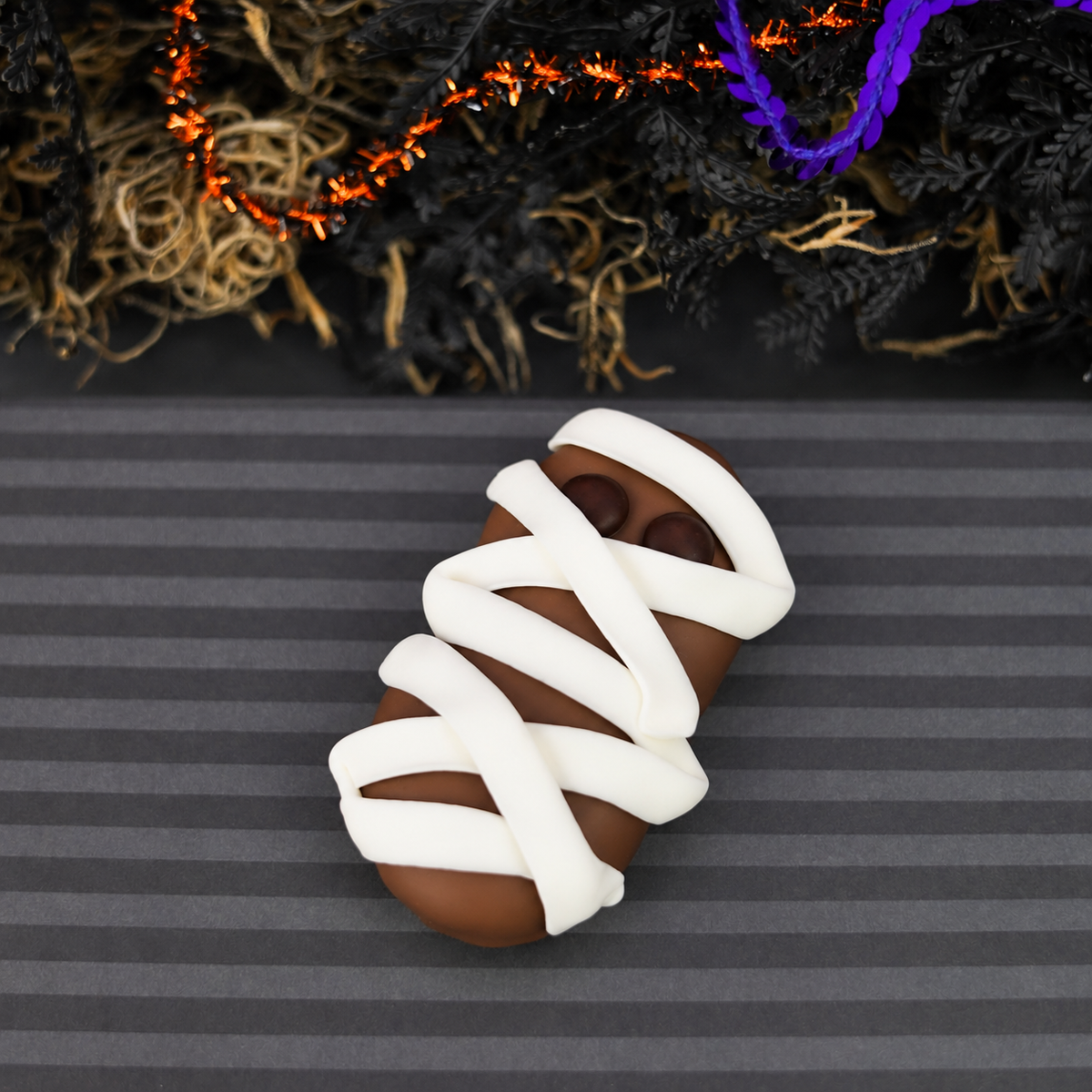 A Halloween-themed mummy nutterbutter cookie, wrapped in white icing with small dark eyes, displayed on a dark gray striped surface with spooky black and orange garlands in the background.