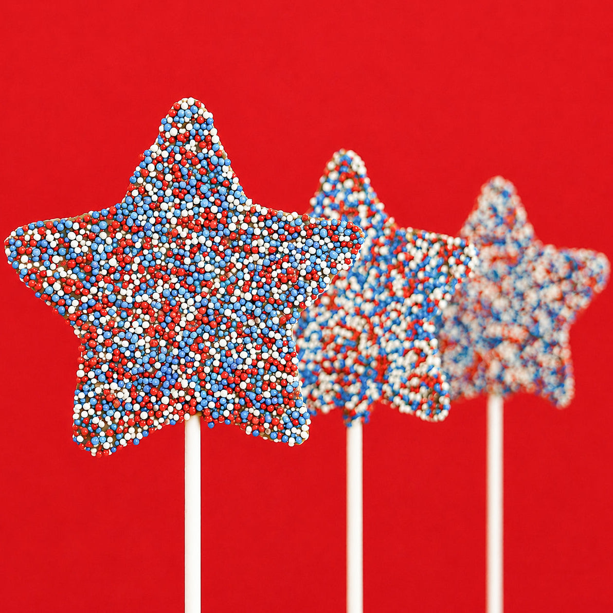 Three solid milk chocolate star lollipops covered entirely in red, white and blue nonpareil candy beads on white sticks against a bold red background, patriotic Fourth of July treat by Nibbles and Bits Scranton PA.