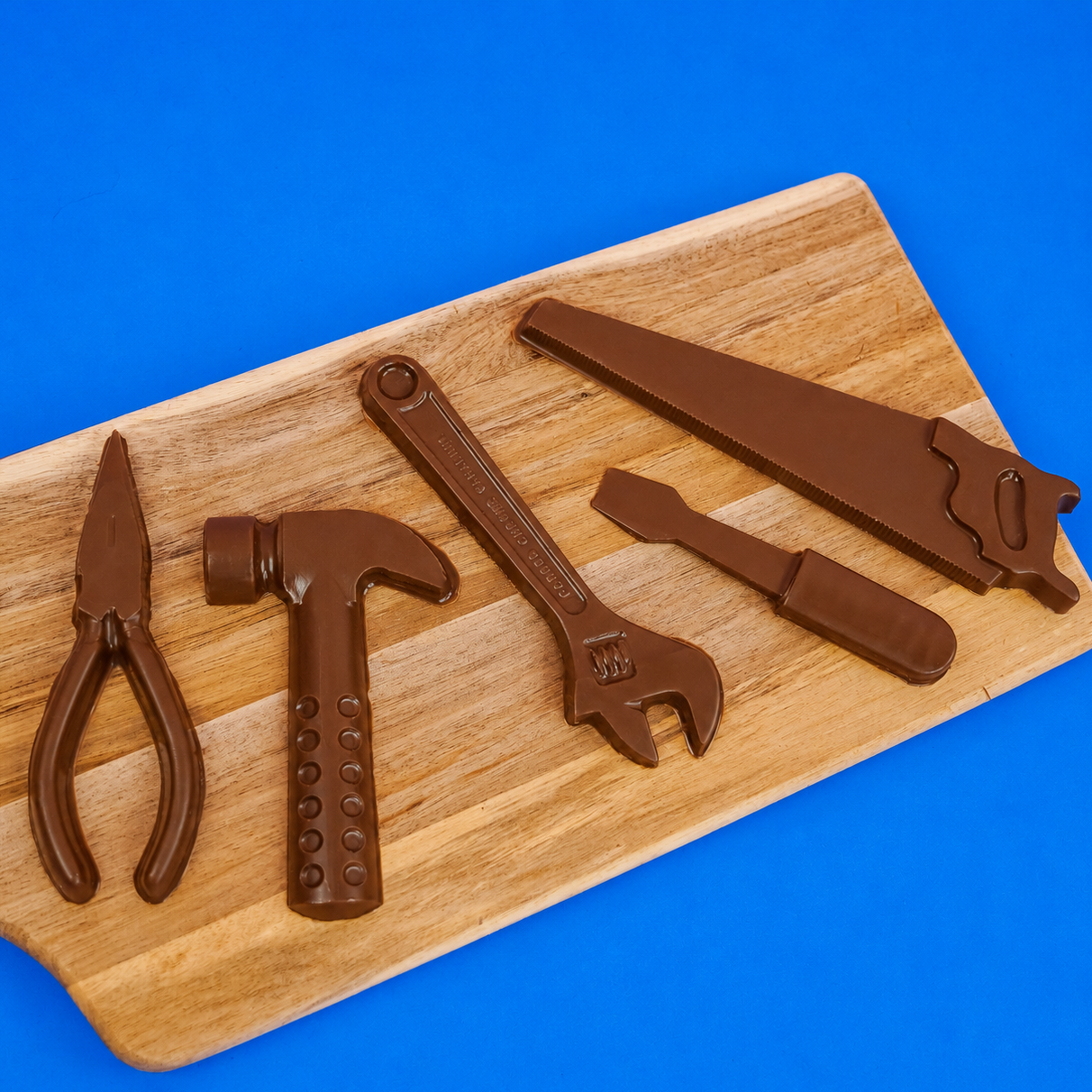 Five small solid milk chocolate tools including pliers hammer wrench screwdriver and hand saw displayed on a rustic wood board with blue background, handmade novelty Father's Day gift by Nibbles and Bits Scranton PA.