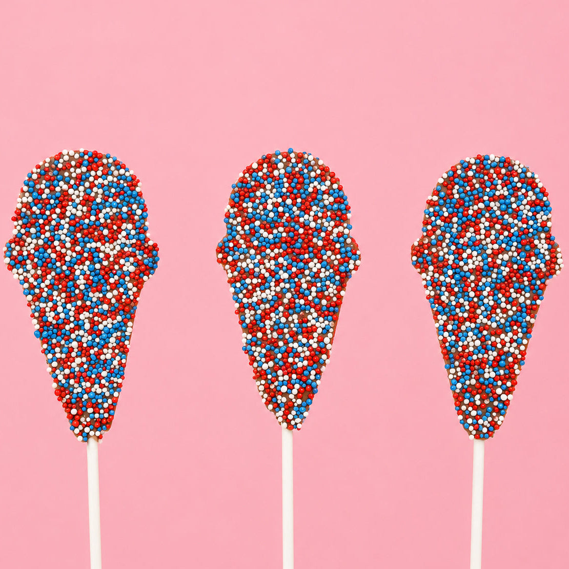 Three solid milk chocolate ice cream cone pops covered entirely in red, white and blue nonpareil beads on white sticks against a pink background, patriotic Fourth of July treat by Nibbles and Bits Scranton PA.