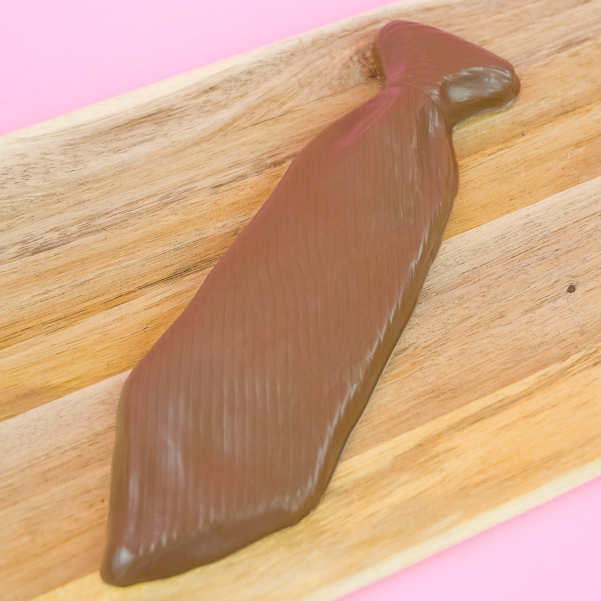 Solid milk chocolate necktie with diagonal stripe texture and molded knot displayed on a rustic wood board with pink background, handmade Father's Day gift by Nibbles and Bits Scranton PA.