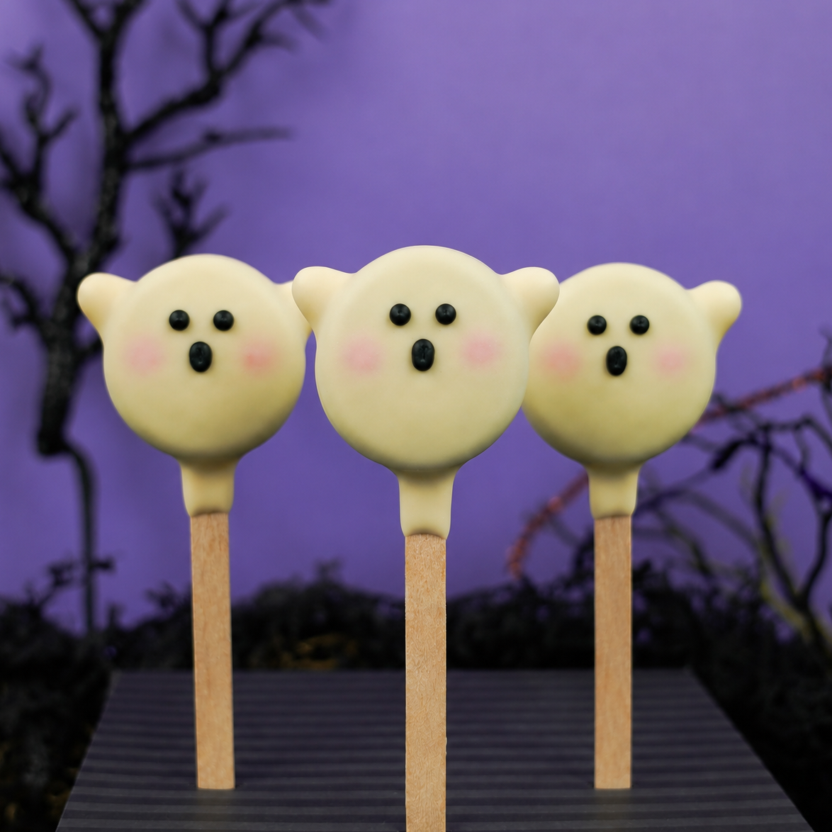 Three white chocolate Ghost Oreo Pops on sticks, hand-painted with ghost faces, blushing cheeks, and mini candy corn ghost arms, against a purple background.