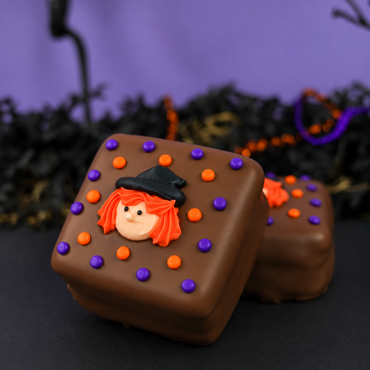 Two chocolate-covered s'mores decorated with orange and purple icing dots and a small witch face topper, photographed on a dark surface with a purple and black Halloween-themed background.