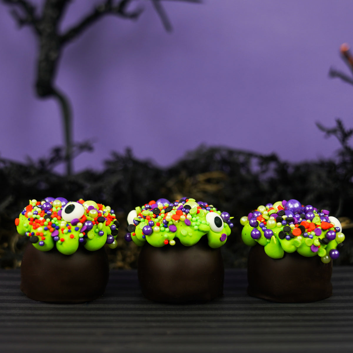 Three dark chocolate cauldron truffles decorated with green goo frosting, halloween sprinkles and eyeballs, arranged on a dark striped surface with a spooky black background.