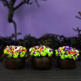 Three dark chocolate cauldron truffles decorated with green goo frosting, halloween sprinkles and eyeballs, arranged on a dark striped surface with a spooky black background.