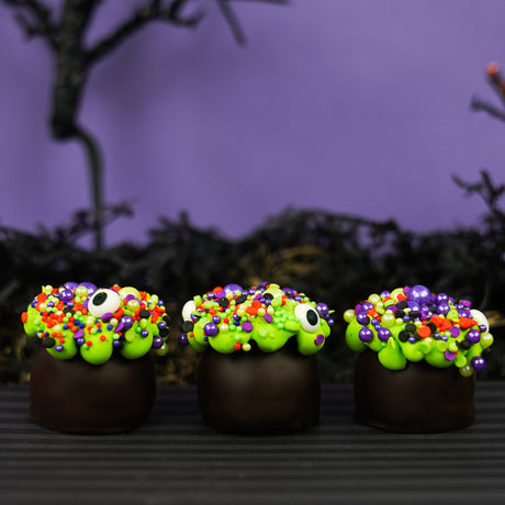 Three dark chocolate cauldron truffles decorated with green goo frosting, halloween sprinkles and eyeballs, arranged on a dark striped surface with a spooky black background.