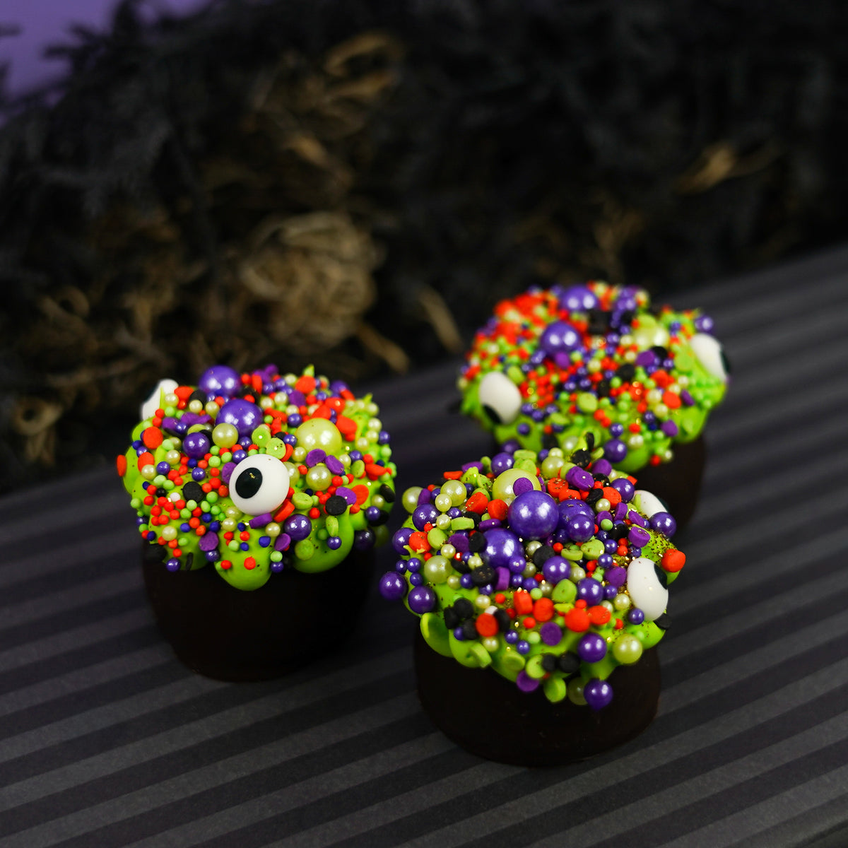Three dark chocolate cauldron truffles decorated with green goo frosting, halloween sprinkles and eyeballs, arranged on a dark striped surface with a spooky black background.