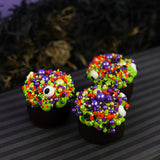 Three dark chocolate cauldron truffles decorated with green goo frosting, halloween sprinkles and eyeballs, arranged on a dark striped surface with a spooky black background.