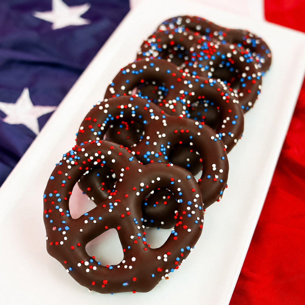Stack of dark chocolate dipped Snyder's pretzels covered in red, white and blue nonpareil candy beads on a white rectangular plate with American flag backdrop, patriotic Fourth of July treat by Nibbles and Bits Scranton PA.