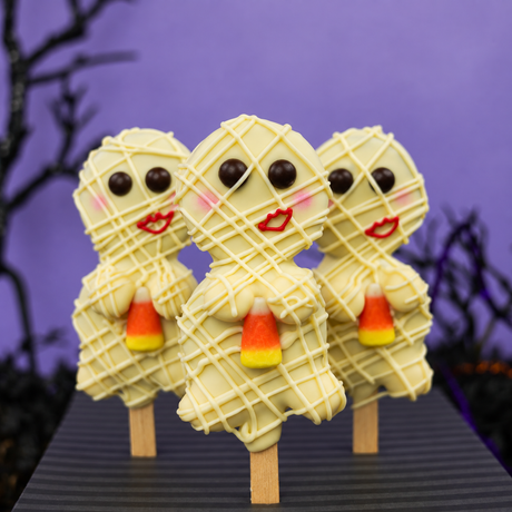 Three Halloween-themed chocolate oreo pops in the shape of mummies, decorated with white chocolate wrap, red lips, and candy corn, displayed on a dark table against a purple background with spooky tree silhouettes.
