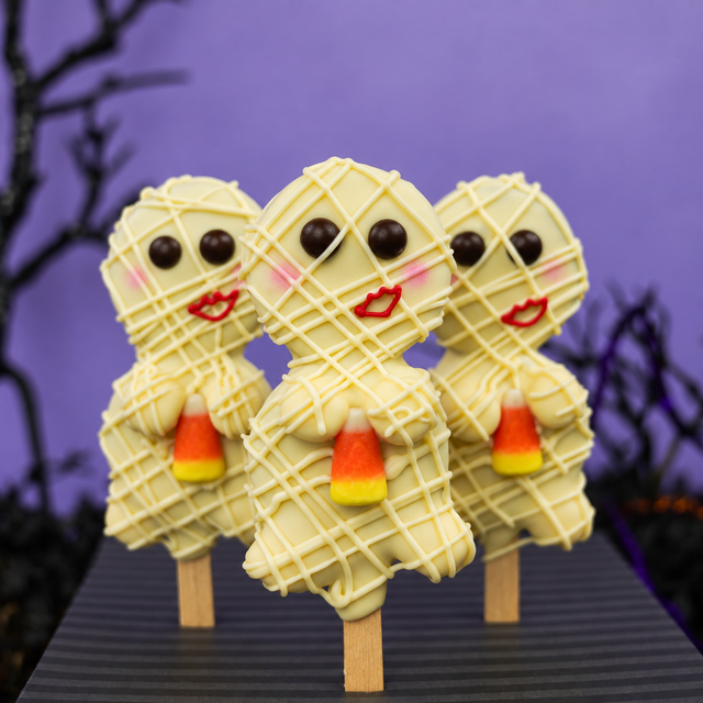 Three Halloween-themed chocolate oreo pops in the shape of mummies, decorated with white chocolate wrap, red lips, and candy corn, displayed on a dark table against a purple background with spooky tree silhouettes.