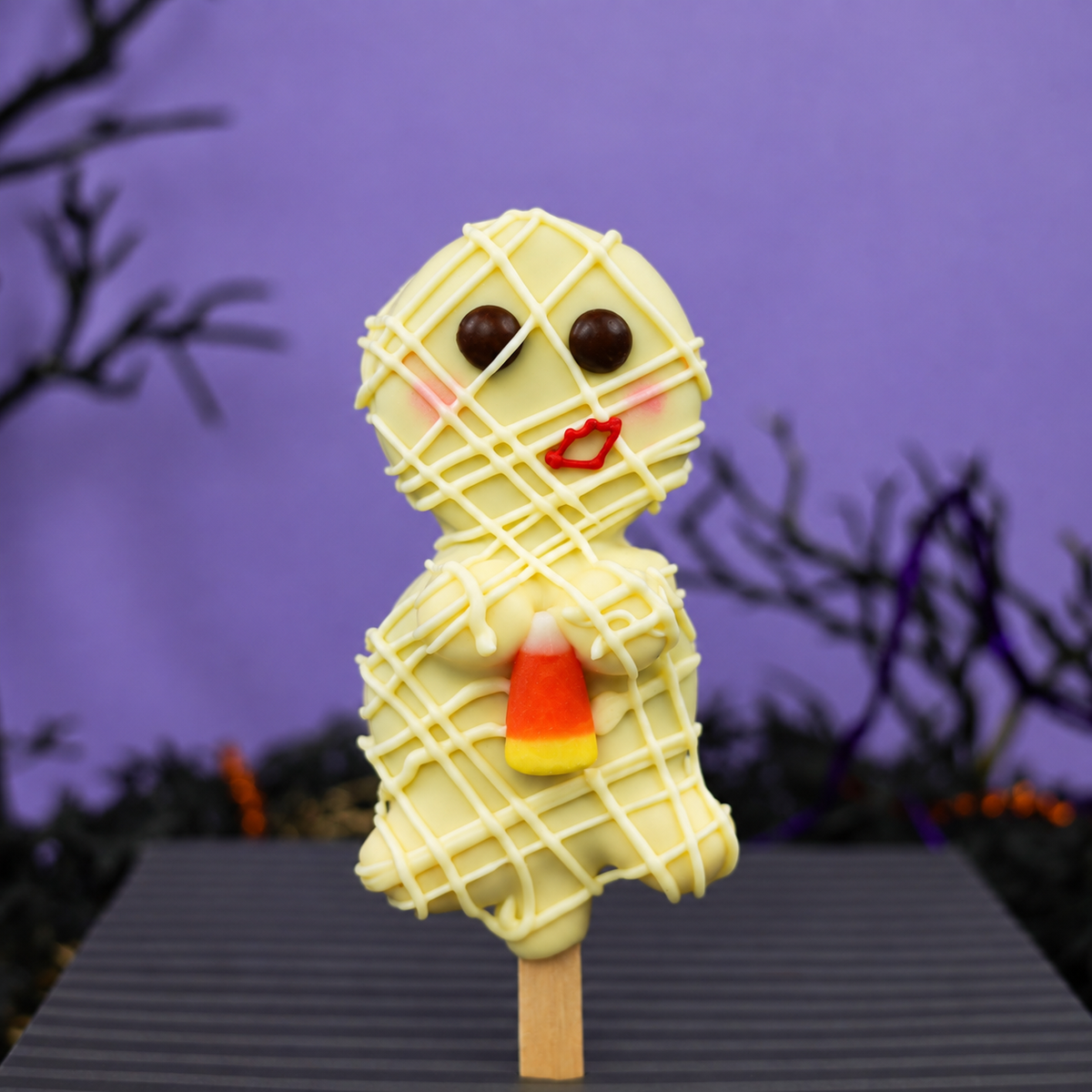 A Halloween-themed white chocolate mummy oreo pop with a candy corn accent, set against a purple background with spooky branches.