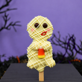 A Halloween-themed white chocolate mummy oreo pop with a candy corn accent, set against a purple background with spooky branches.