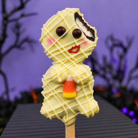 A mummy-shaped oreo pop on a wooden stick, decorated with white chocolate drizzle, chocolate eyes, pink cheeks, red lips, and a bite taken out of a chocolate sandwich cookie on the head. It holds a candy corn in its hands against a purple Halloween-themed background.