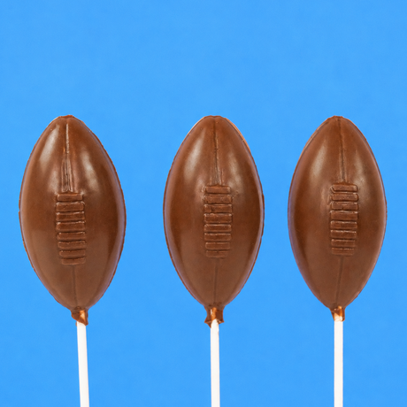 Three solid milk chocolate football shaped lollipops with molded lace stitching detail on white sticks against a blue background, handmade game day treat by Nibbles and Bits Scranton PA.