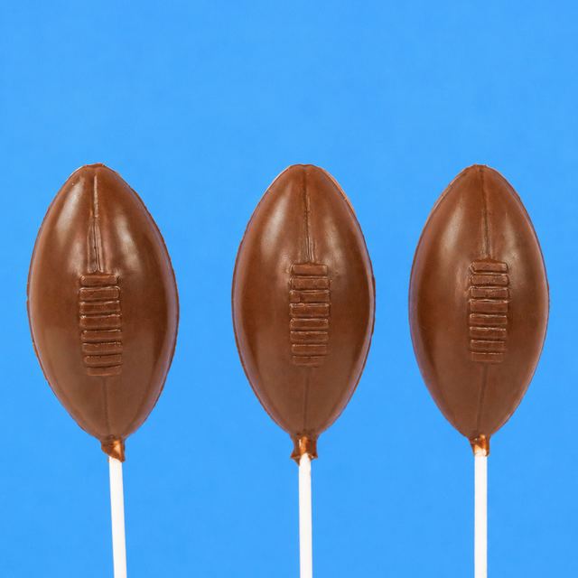 Three solid milk chocolate football shaped lollipops with molded lace stitching detail on white sticks against a blue background, handmade game day treat by Nibbles and Bits Scranton PA.