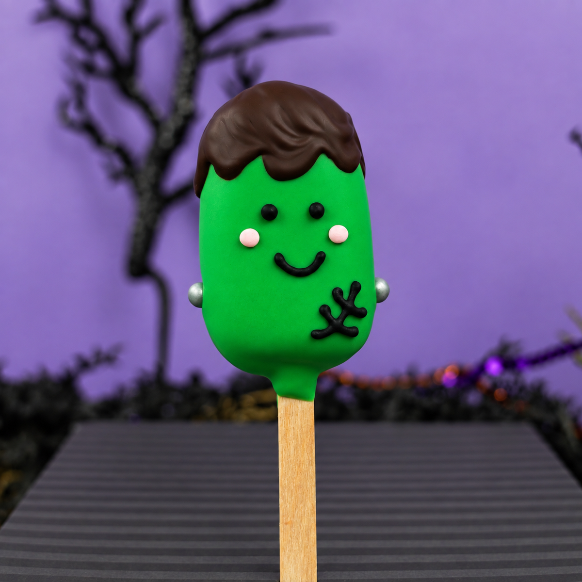 A cookie dough pop dipped in green chocolate and decorated like Frankenstein on a purple Halloween background. 