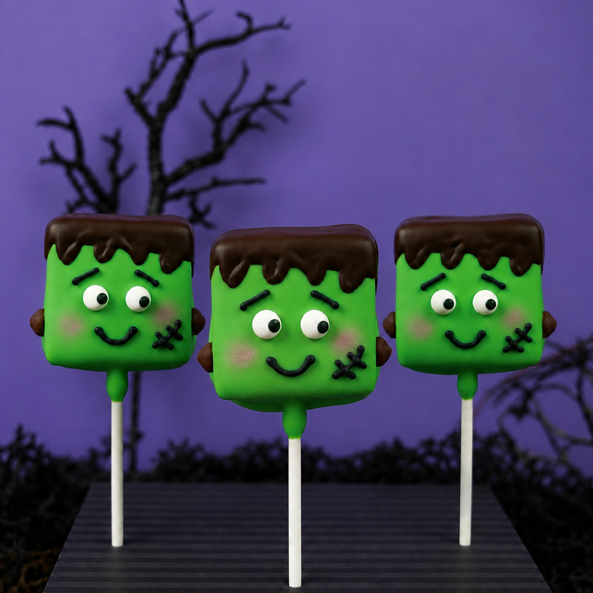 Three Frankie Mallow Pops in a row showing hand-piped stitches, chocolate hair, and candy button eyes, each on a white stick.