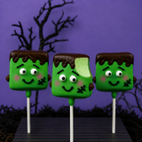 Three green Frankenstein marshmallow pops on a striped tray, one with a bite taken out revealing green marshmallow filling, chocolate hair, and candy eyes against a purple background.