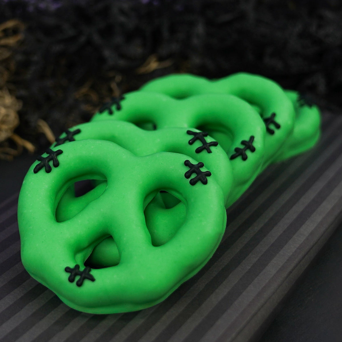 Four bright green chocolate pretzels with black stitched details, arranged on a dark striped surface with a purple and black decorative background.