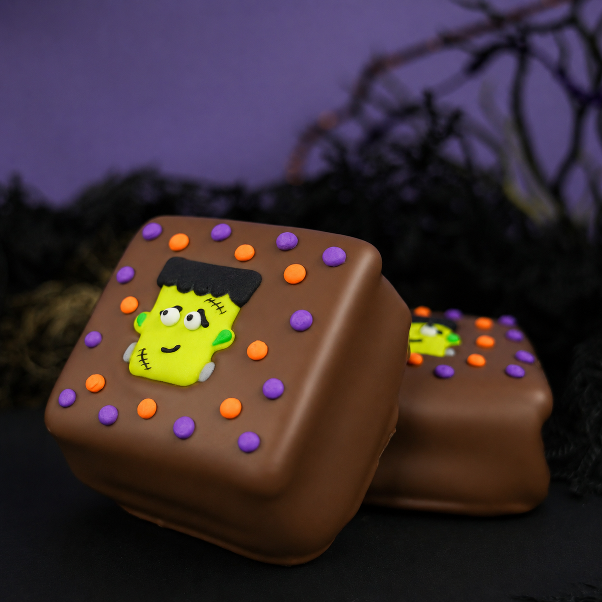 Two Milk Chocolate Frankie S'mores with hand-piped Frankenstein faces and rings of orange and purple icing dots on a dark background.