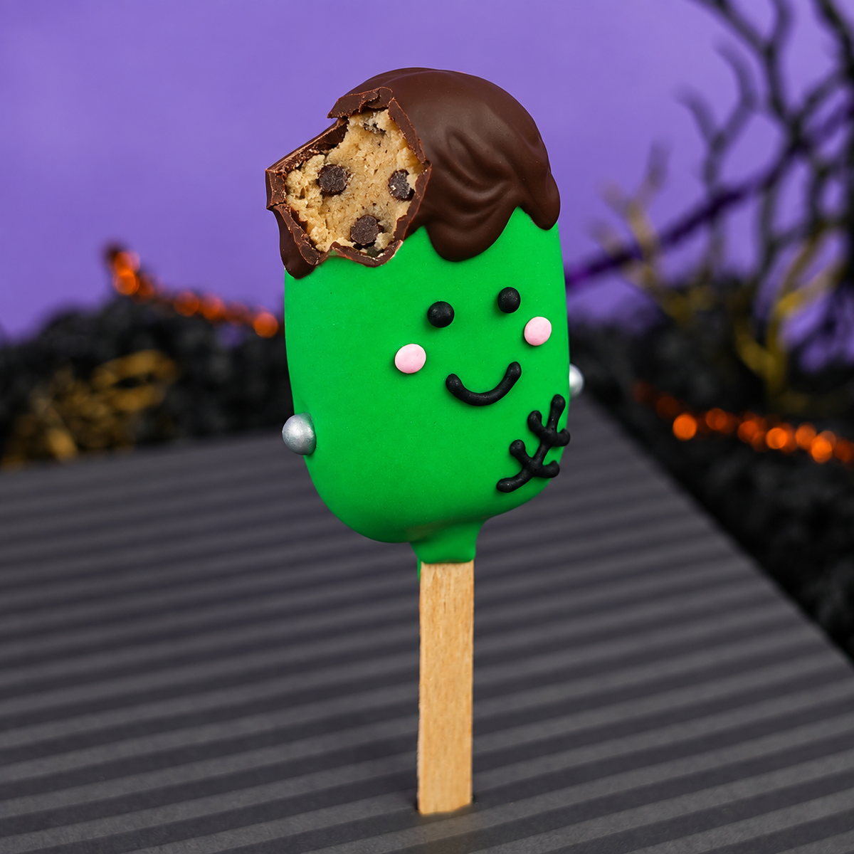 Cookie dough pop dipped in green chocolate and decorated like Frankenstein, with a cut open part at the top to show the cookie dough filling, on a Halloween background. 
