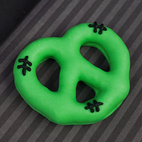 A bright green chocolate pretzel with small black stitched details, photographed on a dark gray striped surface.