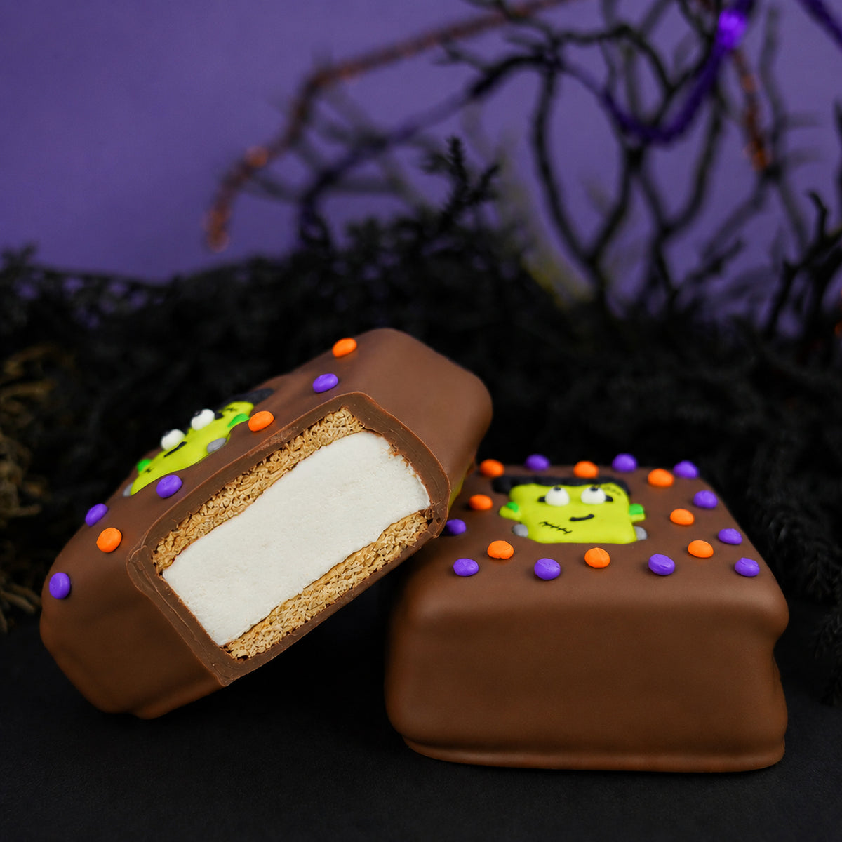 Milk Chocolate Frankie S'more cut in half showing graham cracker and marshmallow layers inside chocolate shell, next to a whole s'more decorated with a Frankenstein face and Halloween icing dots.
