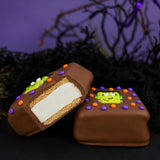 Milk Chocolate Frankie S'more cut in half showing graham cracker and marshmallow layers inside chocolate shell, next to a whole s'more decorated with a Frankenstein face and Halloween icing dots.