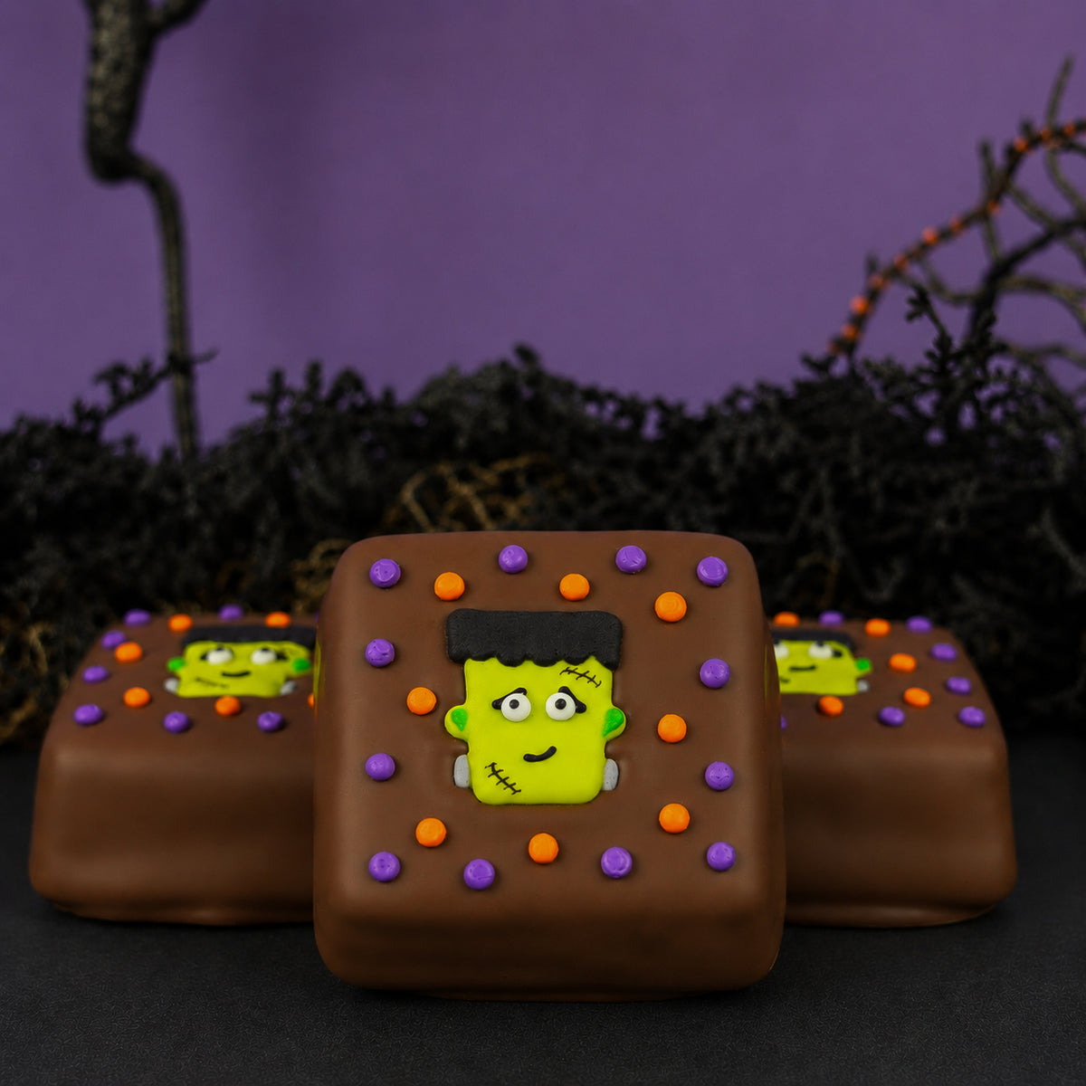 Three Milk Chocolate Frankie S'mores with hand-piped Frankenstein faces and rings of orange and purple icing dots on a dark background.