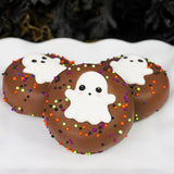 Three milk chocolate Ghost Oreos on a white scalloped plate, each decorated with a hand-piped ghost face and colorful Halloween sprinkles.
