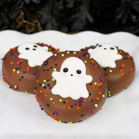 Three milk chocolate Ghost Oreos on a white scalloped plate, each decorated with a hand-piped ghost face and colorful Halloween sprinkles.