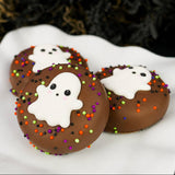 Three milk chocolate Ghost Oreos on a white scalloped plate, each decorated with a hand-piped ghost face and colorful Halloween sprinkles.