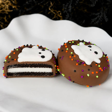 Ghost Oreo cut in half showing the Oreo cookie layers inside milk chocolate shell, next to a whole Ghost Oreo decorated with a white ghost and Halloween nonpareils.