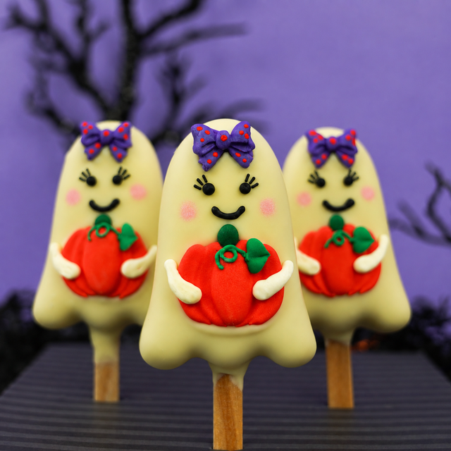 Three Halloween ghost peanut butter pops decorated with purple bows, pink cheeks, and orange pumpkins, set against a purple background with silhouettes of bare trees.