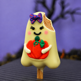 A Halloween ghost peanut butter pop with a bite taken out of the top right, decorated with a purple bow, pink cheeks, and holding a pumpkin. The peanut butter is coated in white chocolate and stands on a wooden stick against a purple background.