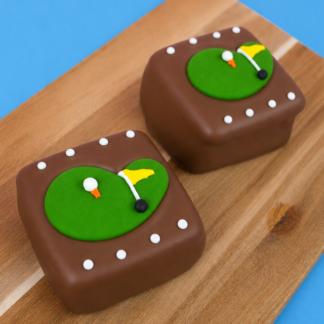 Two milk chocolate golf green s'mores on a rustic wood board with blue background, each decorated with a detailed golf putting green featuring a white golf ball on an orange tee yellow flag black hole and white icing dot details, handmade golf gift by Nibbles and Bits Scranton PA.