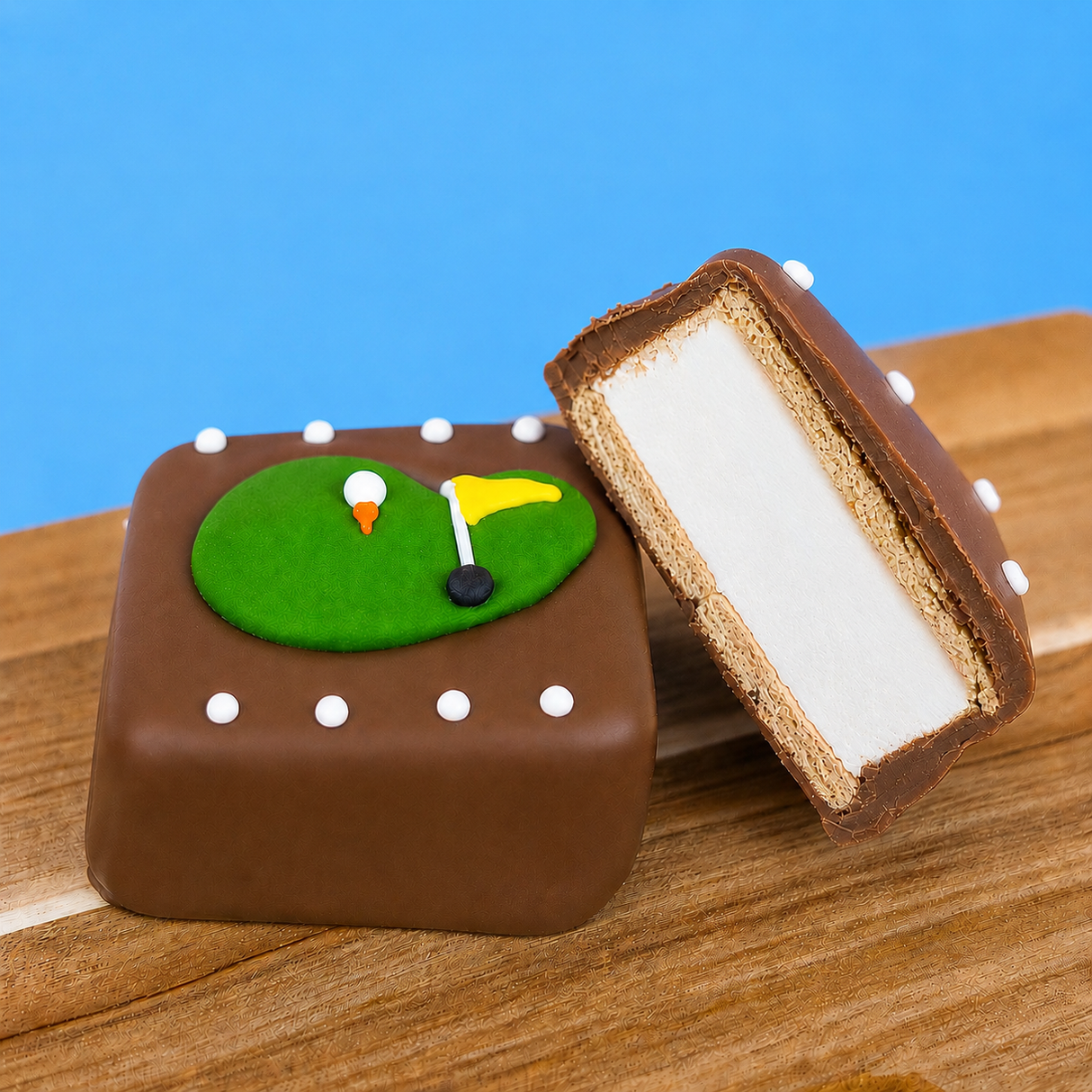 A milk chocolate golf green s'more revealing thick homemade vanilla marshmallow layer and golden graham cracker base alongside a whole decorated s'more on a wood board with blue background, handcrafted golf gift by Nibbles and Bits Scranton PA.